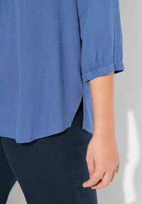 Camicia leggera, blu, a maniche corte con risvolti, spacchi laterali e vestibilità rilassata. Abbinata a jeans scuri. Tessuto liscio.