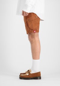 Shorts cargo marron avec poches latérales, ourlet roulé et étiquette rouge. Associés à des mocassins en daim marron et des chaussettes blanches à mi-mollet.