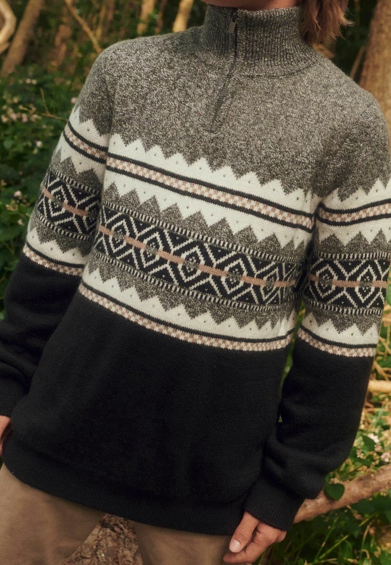 Pull avec col demi-zip en gris, noir et blanc avec des motifs géométriques et en zigzag sur la poitrine et les manches, porté en extérieur.