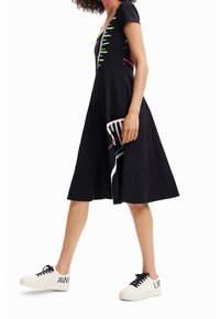 Černé šaty s krátkým rukávem ve tvaru písmene V, zdobené barevnými abstraktními vzory. V kombinaci s bílými teniskami s výrazným textem.