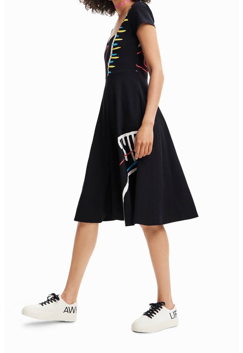 Černé šaty s krátkým rukávem ve tvaru písmene V, zdobené barevnými abstraktními vzory. V kombinaci s bílými teniskami s výrazným textem.