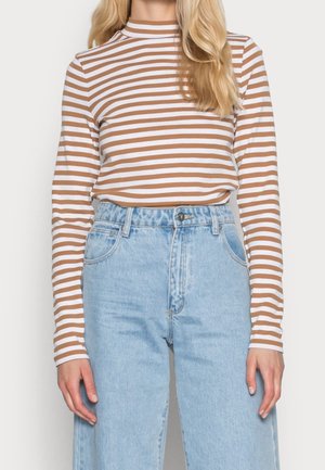Vrouw draagt een bruin-wit gestreepte top met lange mouwen, ingestopt in een lichtblauwe spijkerbroek met hoge taille, staand tegen een witte achtergrond.