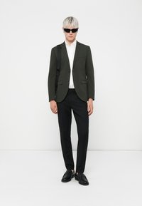 Mørkegrøn blazer over en hvid skjorte, parret med sorte bukser og sko. Modellen bærer sorte solbriller og har en sort taske.