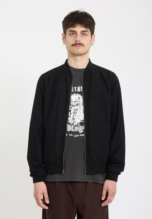Homme avec moustache portant une veste zippée noire par-dessus un t-shirt à motif et un pantalon marron, debout devant un fond blanc uni.