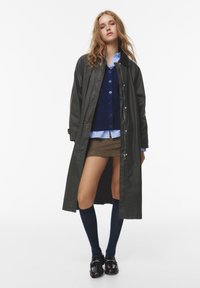 Cappotto nero lungo, cardigan blu navy, camicia bianca a righe, gonna marrone, calze blu fino al ginocchio, scarpe nere. Presenta bottoni e zip.
