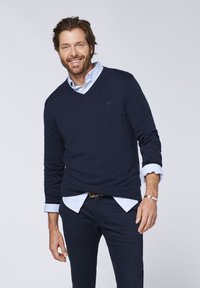 Marineblauer V-Ausschnitt-Pullover aus weichem Strickmaterial, getragen über einem hellblauen Hemd mit Kragen. Hat gerippte Bündchen und ein dezentes Markenlogo.