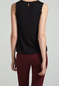 Černý bezrukávový top se scalloped okraji a detailem klíčové díry na zádech, vyrobený z měkké tkaniny. Spárováno s vínovými úzkými kalhotami.
