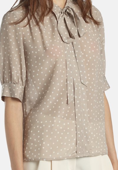 Svetlo bež bluza iz prosojnega blaga z belimi pikami, z ovratnikom z mašnico in kratkimi, zavihanimi rokavi.