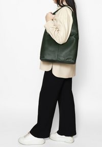 Groene leren draagtas met een gladde textuur, met een brede, ronde vorm en een enkele schouderband, gedragen door een persoon in casual kleding.