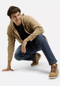 Beżowa bluza z kapturem zapinana na zamek, na czarnej koszulce, w połączeniu z niebieskimi dżinsami i brązowymi wysokimi butami. Model czai się w zrelaksowanej pozie.