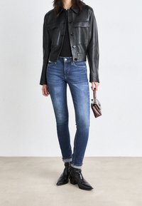 Chaqueta de cuero negra corta con dos bolsillos en el pecho, blusa negra ajustada, jeans ajustados azules y botines negros. Pequeño bolso marrón.