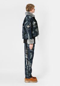 Chupa bomber de mezclilla con cuello de pelaje sintético gris, patrón de camuflaje azul-gris, y múltiples parches; combinado con pantalones de camuflaje a juego y botas color arena.