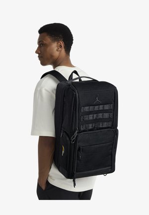 Black backpack with textured fabric, multiple compartments, and adjustable straps. Features a logo and small patch detail on the side.