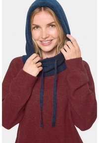 Usmievajúca sa žena v bordovom svetri, ktorá si upravuje tmavomodrú pletenú kapucňu so šnúrkami na jednoduchej pozadí.