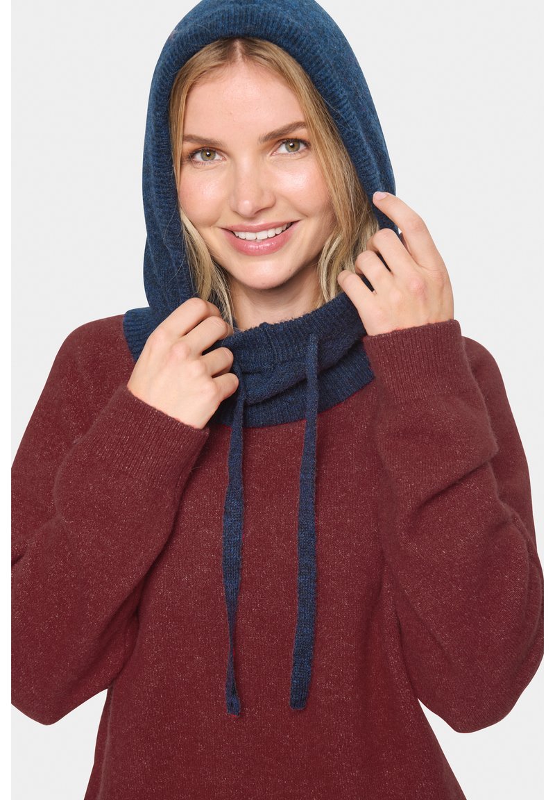 Usmievajúca sa žena v bordovom svetri, ktorá si upravuje tmavomodrú pletenú kapucňu so šnúrkami na jednoduchej pozadí.