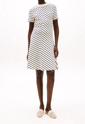 Femme portant une robe blanche à manches courtes et longueur genou avec des rayures diagonales noires et des chaussures blanches tissées sans lacets, debout sur un fond blanc.