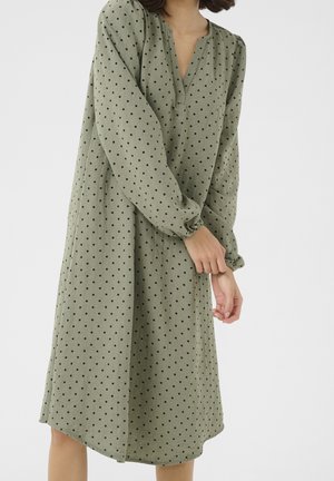 Femme portant une robe ample, vert olive, à manches longues avec des pois noirs, ajustant le poignet de la manche, sur un fond clair uni.