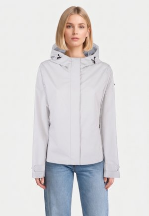 Donna che indossa una giacca con cappuccio grigio chiaro con cordini e jeans blu, in piedi contro uno sfondo bianco uniforme.