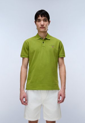 Mężczyzna stoi zwrócony przodem, ubrany w zieloną koszulkę polo i białe spodenki na jednolitym jasnym tle.