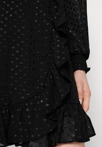Svart klänning med texturerat prickmönster, med volanger längs fållen och ärmarna. Materialet verkar lätt och transparent.