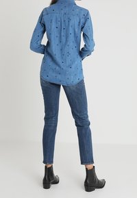 Denim blouse met geborduurde hart- en driehoekpatronen, lange mouwen, een spitse kraag en een losse pasvorm, gedragen met een jeans en enkellaarzen.