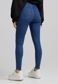 Jeans de mezclilla azul ajustados con cintura alta y bolsillos traseros, combinados con zapatillas blancas. La tela tiene una textura suave.