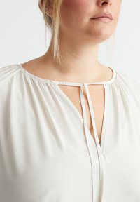 Weiße, fließende Bluse mit einem V-Ausschnitt und gebundenem Akzent, gerafften Schultern und glatter Textur. Geeignet für Freizeit- oder formelle Anlässe.