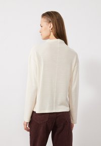 Pull en maille couleur crème avec un col haut et des épaules déchues, présentant une texture lisse et un détail de couture au dos. Porté avec un pantalon bordeaux.