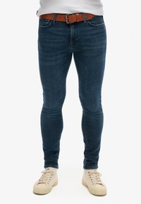 Jeans de denim ajustados en azul oscuro, con una superficie texturizada. Llevados con un cinturón de cuero marrón y zapatillas beige.