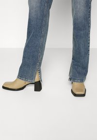Botas de tobillo beige con punta cuadrada y tacón grueso negro, llevadas con jeans azul claro que tienen slit en los laterales. Textura de cuero suave visible.