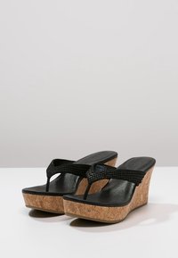 Černé klínové sandály s korekovou základnou, texturovanými černými pásky a hladkým polštářem pro nohu. Disponují plochým designem mezi prsty pro snadné obouvání.