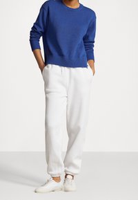 Personne portant un pull bleu en maille, un pantalon de jogging blanc avec poches, et des baskets blanches décontractées, debout les mains dans les poches.