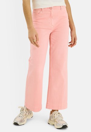 Persona con pantalones rosas de talle alto y pierna ancha con bolsillos frontales y zapatillas beige gruesas, de pie con las manos relajadas a los lados.