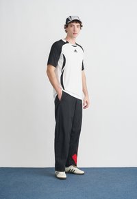 Biele športové tričko s čiernymi prvkami a logom, v kombinácii s čiernymi nohavicami s červeným trojuholníkovým dizajnovým detailom. Bielobiele športové topánky.