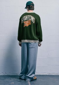 Pullover verde lavorato a maglia con "HAWKINS High" ricamato in arancione e crema, abbinato a pantaloni della tuta grigi con tasca laterale.