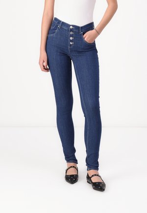 Persoon draagt een donkere blauwe skinny jeans met hoge taille en knoopsluiting, een wit topje en zwarte puntschoentjes met enkelbandjes en oogjes.