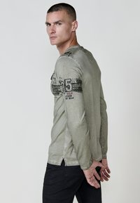 Hosszú ujjú zöld póló grafikai részletekkel és szöveggel az ujjon, fekete nadrággal párosítva. A póló laza fitettel rendelkezik, oldalsó részen hasítékkal.