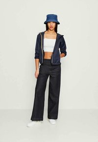 Casaco navy com fecho e capuz, top cropped branco, jeans largos escuros e ténis brancos. Chapéu bucket azul adiciona um detalhe de contraste.