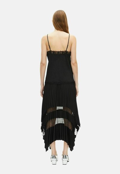 Mujer con cabello largo vistiendo una camiseta de tirantes con encaje negro y una falda plisada negra con paneles de bolitas transparentes, tacones plateados, de pie de espaldas.
