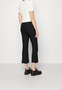 Schwarze, taillierte Cropped-Hosen, kombiniert mit einem strukturierten weißen Strickoberteil. Das Model trägt schwarze Schnürschuhe mit dicken Sohlen.