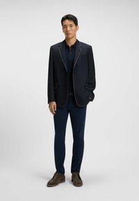Navy blazer with white stitching, paired with a navy shirt and slim-fit pants. Brown shoes complete the outfit, showcasing a tailored look.