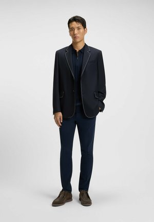 Navy blazer with white stitching, paired with a navy shirt and slim-fit pants. Brown shoes complete the outfit, showcasing a tailored look.
