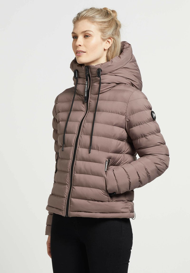 Blondynka w mauve ocieplanej kurtce puffer z wysokim kołnierzem i kapturem, czarnymi sznurkami, z rękami w kieszeniach, zwrócona w prawo.