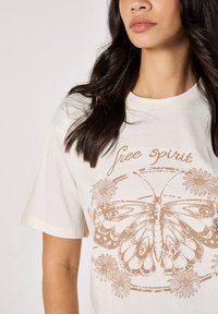 Camiseta de color crema con un gran gráfico de una mariposa en marrón, rodeada de elementos florales y el texto "espíritu libre" arriba.