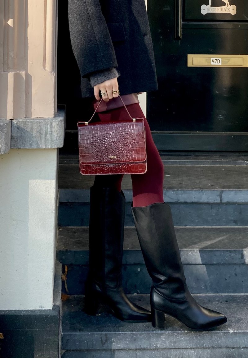 Kleine burgunderfarbene Handtasche aus Kunstleder im Krokodil-Look mit einem oberen Griff, kombiniert mit schwarzen kniehohen Stiefeln mit Absatz und maroonfarbenen Leggings.