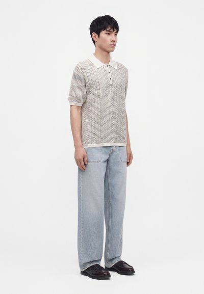 Jeune homme debout portant un polo beige à manches courtes à motif, un jean bleu clair à jambes larges et des chaussures marron foncé sur fond blanc.
