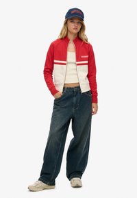 Rood-wit bomberjack met rits, oversized donkere denimbroek en beige sneakers; blauwe pet met "SUPERDRY"-logo.