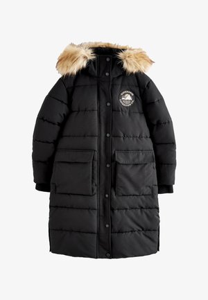 Ung person iført en lang sort dynejakke med pels hætte, beige bukser, hvide sneakers og en brun sweater mod en ensfarvet baggrund.