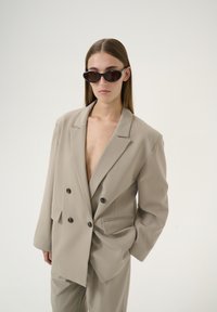 Beige, zweireihiger Blazer aus strukturiertem Stoff mit großen schwarzen Knöpfen, kombiniert mit passender Hose und schmalen schwarzen Sonnenbrillen.