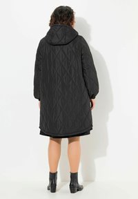 Cappotto quiltato nero con cappuccio, caratterizzato da una silhouette lunga, motivo a rombi e polsini elasticizzati. Indossato sopra un abito scuro e stivaletti alla caviglia.